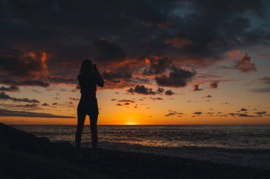 Deniz kıyısında duran ve güzel, renkli, sihirli bir günbatımını cep telefonuyla çeken narin bir kadının silueti.