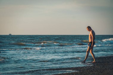 Yüzme şortlu mutlu adam gün batımından önce kıyıda duruyor ve film tarzında fotoğraf çekmekten hoşlanıyor. Deniz kenarında tatil.
