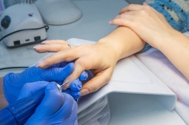 Mavi eldivenli bir manikür ustasının ellerini yakın plan bir güzellik salonundaki tırnak altlarını budamak ve sökmek için elektrikli tırnak törpüsü kullanıyor. Donanım manikürü
