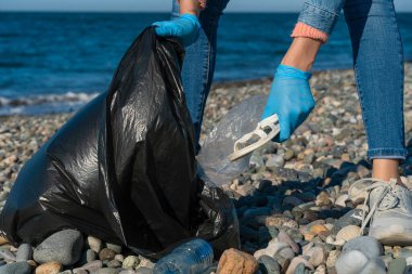 Bir kadının elleri, denizin çakıl taşlı sahilinde siyah bir çöp torbasında plastik topluyor. Karadeniz kıyısındaki çevre kirliliği.