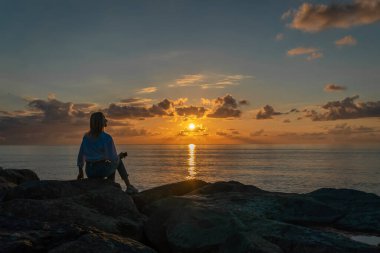 Beyaz gömlekli ve kotlu genç bir kadın büyük kayaların üzerinde oturuyor ve denizin üzerinde büyüleyici bir gün batımına bakıyor, arkadan çekilmiş bir fotoğraf, uzay kopyası.