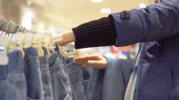 Manos de mujer joven elegir vaqueros que cuelgan de la percha en la tienda de ropa. — Vídeos de Stock