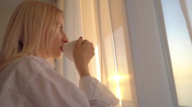 Bir fincan kahve ya da çay içen düşünceli genç bir sarışın pencereden dışarı bakar.,