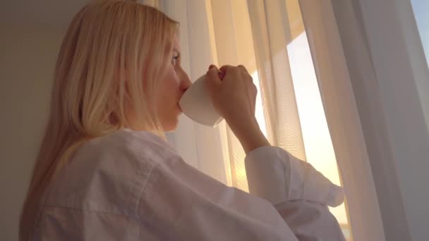 Rubia con una camisa blanca mira por la ventana, bebe una bebida caliente de té o café, — Vídeos de Stock