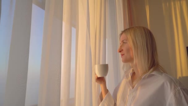 Rubia con una camisa blanca mira por la ventana, bebe una bebida caliente de té o café, — Vídeos de Stock