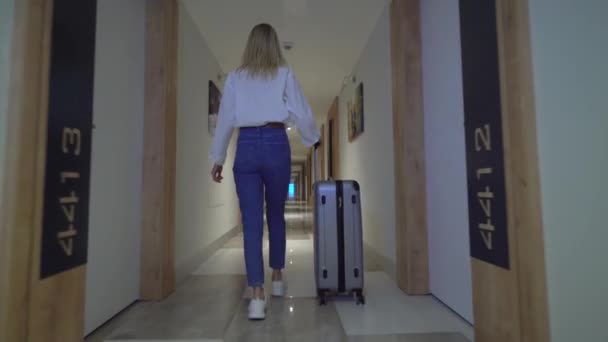 Вид сзади на женщину, идущую по коридору отеля. закатывает багаж за спину. — стоковое видео