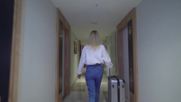 Kadın otelin koridorunda yürüyor ve bavulunu tekerlekli bavulla yuvarlıyor.. — Stok video