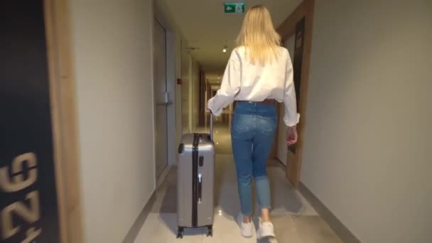 Kadın otelin koridorunda yürüyor ve bavulunu tekerlekli bavulla yuvarlıyor.. — Stok video
