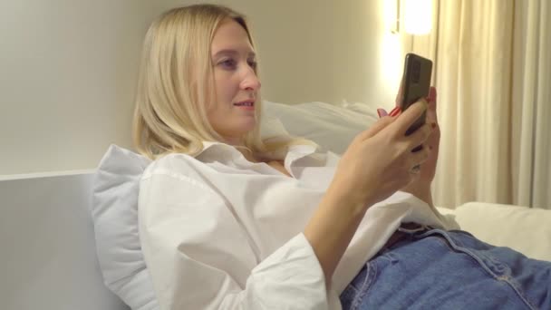 A woman at home reads a text message on her smartphone while lying on the bed. — Stock Video