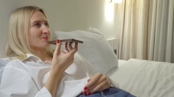 Žena doma nahrává hlasovou zprávu na svém smartphonu, zatímco leží na posteli. — Stock video