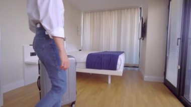 Turist bavuluyla apartmana giriyor, perdeleri itiyor ve manzaranın tadını çıkarıyor.