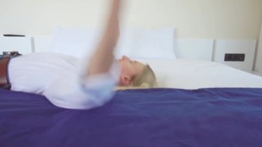 Beyaz gömlekli bir sarışın ağır çekimde beyaz bir yatağa düşüyor..
