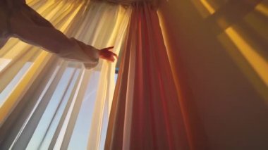 Kadın perdeleri açar ve güneşin doğuşunu seyreder. Gün batımı ışınları siluete düşer.