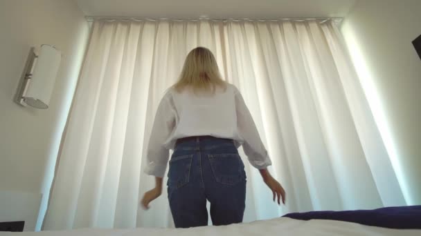 Beyaz gömlekli bir kadın yataktan çıkar ve penceredeki perdeleri açar.. — Stok video