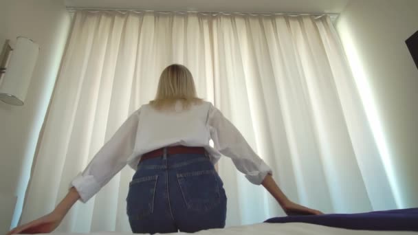 Een vrouw in een wit shirt komt uit bed en doet de gordijnen van het raam open. — Stockvideo