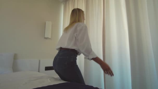 Una mujer con una camisa blanca se levanta de la cama y abre las cortinas de la ventana. — Vídeos de Stock