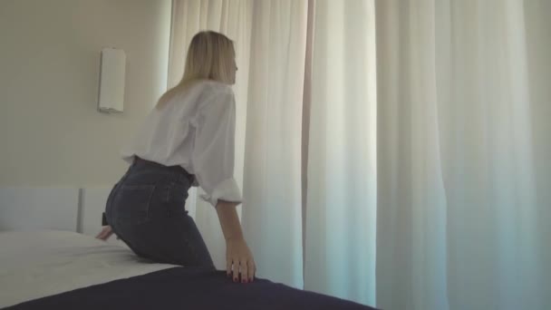 En kvinna i vit skjorta kliver upp ur sängen och öppnar gardinerna på fönstret. — Stockvideo
