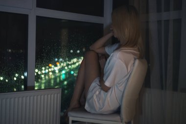 Beyaz gömlekli genç sarışın kadın çenesi yukarıda bir sandalyede oturur ve bir yerden bir tavana gece şehrini seyreder.