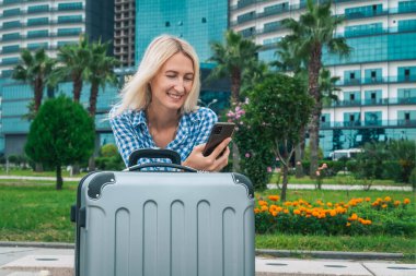 Mavi ekose gömlekli, elinde cep telefonu olan genç, sırıtan sarışın bir kadın ve bir bavul yeşil çalıların arkasında duruyor.