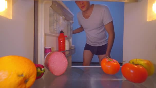 İç çamaşırlı adam buzdolabını açıyor, esniyor, ağzını kapatıyor, domatesi alıyor. — Stok video