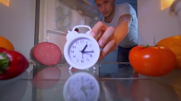 Молодой человек или парень открывает холодильник, у которого есть будильник, он берет его — стоковое видео