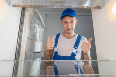 Tulumlu gülümseyen erkek teknisyen baş parmağını kaldırmış. Buzdolabı tamir konsepti. Buzdolabının içinden fotoğraf.
