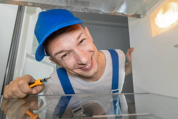 Portrait of a young smiling male worker in uniform inside a refrigerator with a screwdriver. Concept of refrigerator repair, hired work. Photos inside the refrigerator