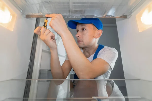 Male technician in overalls repairs a broken refrigerator with a screwdriver. Refrigerator repair concept. Photo from inside the refrigerator