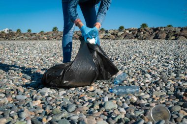 Kadın güneşli bir günde, çakıl taşı plajında plastik, köpük ve kauçuk topluyor. Kıyı bölgesinin temizlenmesi. Ekoloji konsepti, gönüllülük.