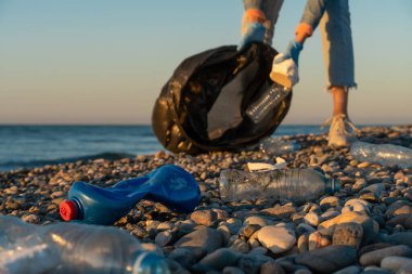 Gün batımında deniz çakıl taşı plajında siyah çöp torbası taşıyan genç bir kadının arka planına yakın çekim. Ekoloji, gönüllü olma, çevre kirliliği kavramı.