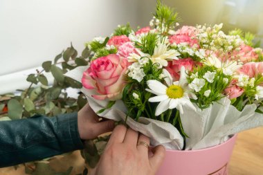 Genç bir kadın çiçekçinin ellerinin yakın çekimi masanın üzerindeki karton kutuya ve kağıda narin pembe güller, karanfiller ve beyaz papatyalardan oluşan güzel bir kompozisyon yerleştiriyor..