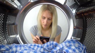 Kadın ev hanımı akıllı telefonuna çamaşır makinesinin açık kapısından bakar. Davulun içinden bak. Giysiler. Çamaşır makinesi arızası ya da uygulama hatası. Garanti ya da onarım.