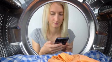 Genç, güzel, ince, beyaz bir kız çamaşır makinesinin önünde oturuyor, telefonu elinde tutuyor ve içine bakıyor. Başvuru ya da talimat, yıkama kılavuzu. Kamera içeriden geniş açıyla çeker
