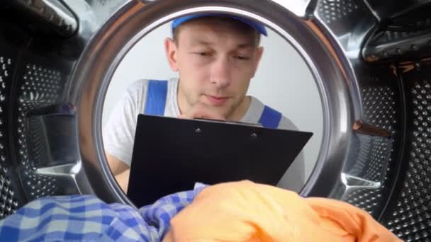 若い洗濯機修理工はドラムを見て ペンでタブレットで作業のリストを書き込みます 彼は青い制服を着ている 家電の修理 広い角度から見ると — ストック動画
