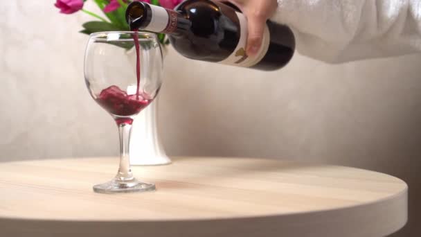 白いコートを着た女性の手は ボトルから赤ワインをテーブルの上に立つガラスに注ぐ アパートでワインの試飲 サムリエ 病気としての飲酒 — ストック動画