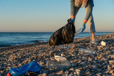 Genç bir kadın gün batımında deniz kıyısındaki bir çakıl taşlı sahilde siyah bir çöp torbasında plastik topluyor. Kullanılmış kirli plastik şişeler. Karadeniz kıyısındaki çevre kirliliği