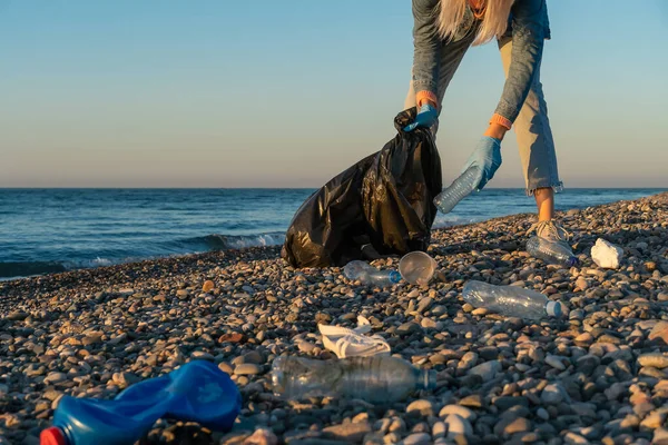 Young woman collects plastic, rubber garbage in a black garbage bag on a pebble beach by the sea at sunset. Empty used dirty plastic bottles. Environmental pollution on the Black Sea coast