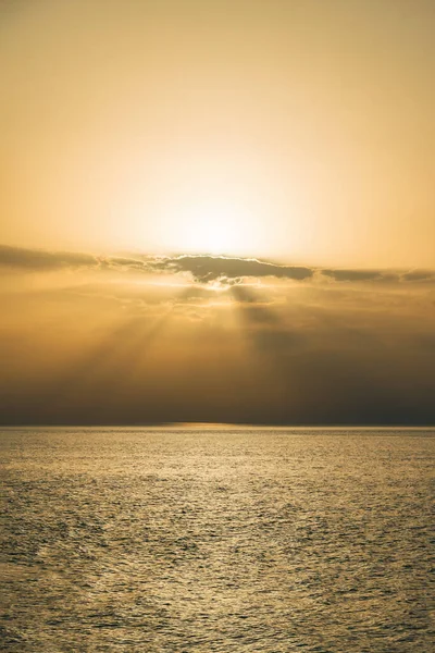 Yellow-orange sun has hidden behind clouds over the sea, the sun is releasing rays from under the clouds. Beautiful seascape with the sun. Vertical photo