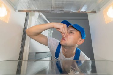 Özel iş elbiseleri içindeki erkek teknisyen buzdolabındaki nahoş koku yüzünden burnunu parmaklarıyla kapadı. Bozuk buzdolabı kavramı, tamir işi. Buzdolabının içinden fotoğraf.