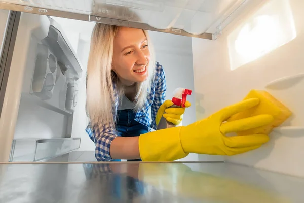 View from inside the refrigerator. Smiling woman in rubber gloves and with a cleaning agent cleans an empty refrigerator with a sponge.