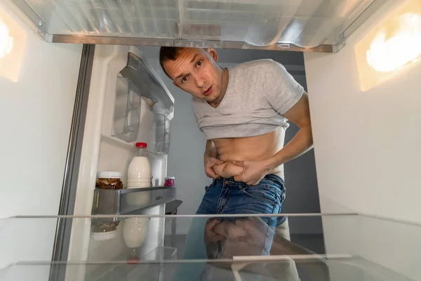 Young man opens the refrigerator, looks inside, where the apple is. He sad takes himself by the fat folds on his stomach and thereby shows that he is on a diet. Weight loss.