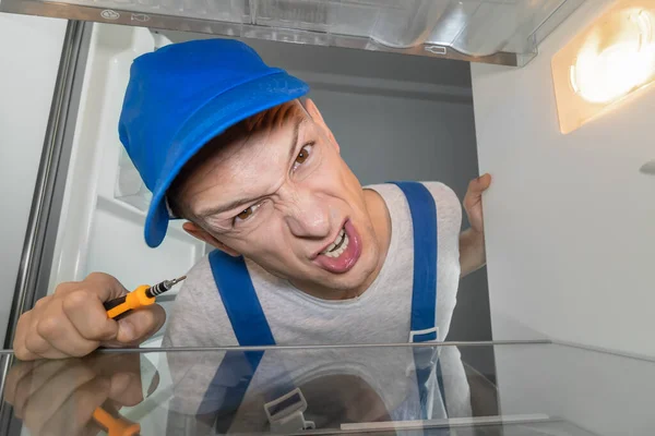 Portrait of a young, grimacing, angry male worker in uniform inside a refrigerator with a screwdriver. Concept of refrigerator repair, hired work. Photos inside the refrigerator