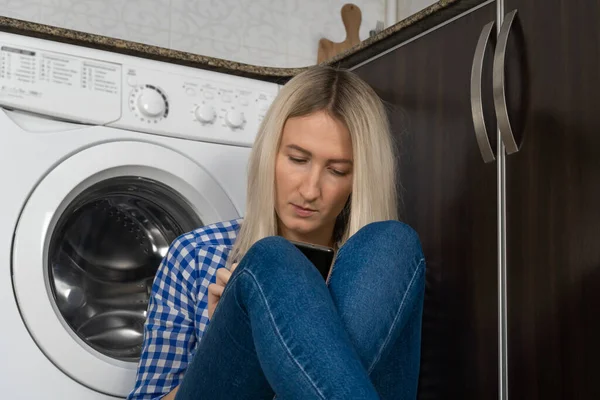 Close up young blonde woman with a serious concentrated look looks at her mobile phone sitting in the kitchen near the washing machine