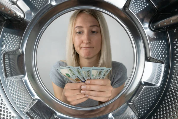 Young woman holds money in her hands near the washing machine and looks with surprise at the side, a photo from inside the drum of the washing machine. Concept of payment, money laundering.