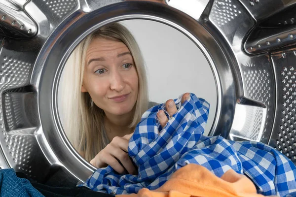 Disgruntled young blonde woman holds a leaky shirt in her hands after washing, photo from inside the drum of a washing machine. Woman is disappointed with the work of the washing machine