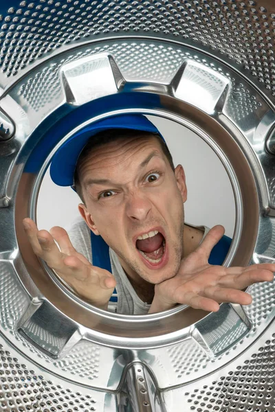 Angry screaming male repairman in uniform looks into the drum of the washing machine, photo from inside. Vertical photo