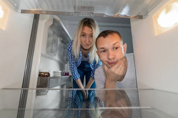 Young man and a woman look into an empty refrigerator. Concept of a delivery service, poverty, crisis. Photo from inside the refrigerator