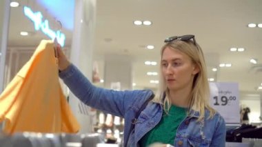Beautiful young Caucasian woman with white hair chooses clothes in store or shopping center. She removes her clothes from hanger and inspects them. shirts and jeans.