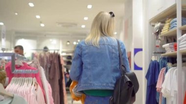 Beyaz saçlı güzel beyaz kadın mağazadan ya da alışveriş merkezinden kıyafet seçiyor. Elbiselerini askıdan çıkarıyor ve onları inceliyor. Gömlek ve kot.