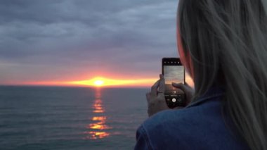 Kadınların elleri yakın plan akıllı telefonu tutar ve üzerinde güzel turuncu gün batımını fotoğraflardı. Bulutlar renkli kırmızıdır. Dikiz aynası. Tatildeki neşeli ruh hali.